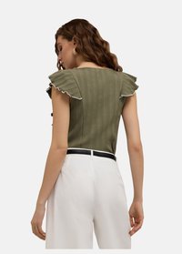 Olivgrön ribbad topp med dekorativa volangärmar och vit kant, matchad med högmidjade vita byxor och ett svart bälte.