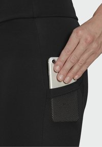 Czarne legginsy sportowe z boczną kieszenią, w której znajduje się srebrny smartfon. Kieszeń ma teksturowany siatkowy wzór z małymi perforacjami.