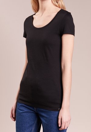 Vrouw die een strakke zwarte T-shirt met korte mouwen en een ronde hals draagt, gecombineerd met een blauwe spijkerbroek, tegen een neutrale achtergrond.