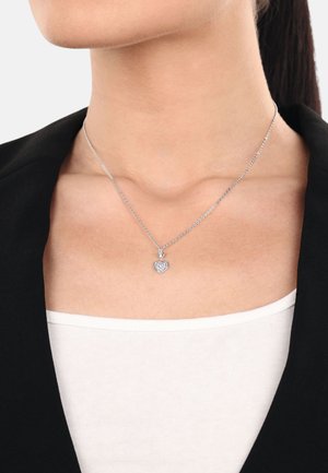 Collana con pendente a forma di cuore in argento, con superficie testurizzata, dotata di un piccolo gemma trasparente, su una delicata catena, indossata sopra una camicia bianca e un blazer nero.