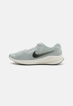 Zapatilla deportiva gris claro con parte superior de malla, acentos negros y suela blanca. Presenta una punta redondeada y un diseño de cordones. Superficie texturizada.
