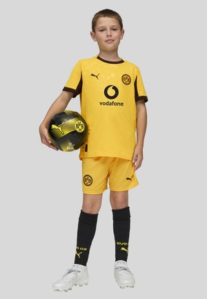 Camiseta de fútbol amarilla con detalles en marrón, logo negro; pantalones cortos a juego; medias negras con ribete amarillo; sosteniendo un balón de fútbol estampado en negro y amarillo.