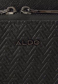 Černá texturovaná kabelka s cik-cak vzorem, stříbrnými doplňky a embosovaným logem "ALDO" na přední straně. Viditelný detail se zipovým uzávěrem.
