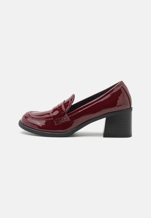Burgundy patent leather loafers with a rounded toe, front strap detail, and a black block heel. Sleek, glossy finish and structured design.