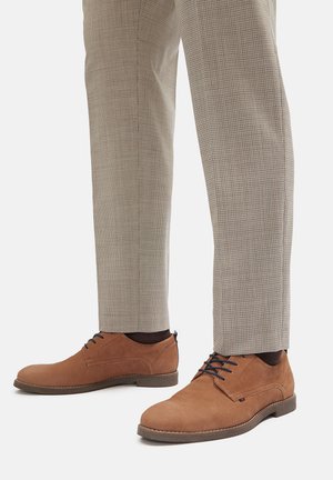 Brązowe zamszowe buty z okrągłym noskiem, wiązane sznurowadłami w kontrastowym ciemnym kolorze. Noszone z jasnymi, kratkowymi spodniami.