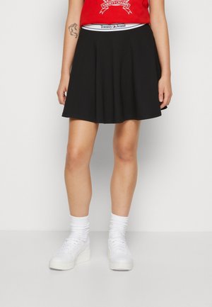 Vrouw die een zwarte Tommy Jeans-rok draagt met een witte tailleband, witte sneakers en witte sokken, staand tegen een effen lichte achtergrond.
