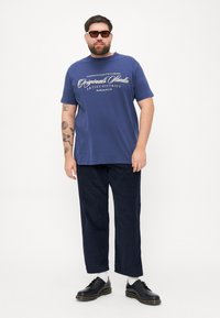 Navy blue cotton t-shirt with white text, relaxed fit, short sleeves, paired with dark navy pants and black lace-up shoes, white socks visible.