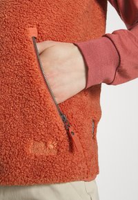 Gilet in fleece arancione con superficie tessurizzata, tasca con zip e logo. Camicia a maniche lunghe sotto in rosa tenue.