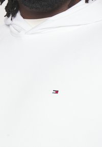 Sudadera blanca hecha de tela suave, que presenta un pequeño logo bordado en rojo, azul marino y blanco en la parte izquierda del pecho.
