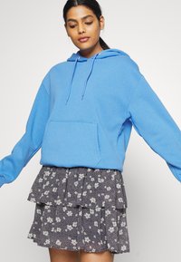 Sweatshirt azul claro com bolso frontal e capuz ajustável, sobre uma saia em camadas com padrão floral azul-marinho e pregas.