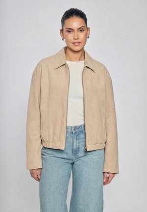Frau mit zurückgebundenem dunklem Haar trägt eine beige Jacke mit Reißverschluss, ein weißes Oberteil und hellblaue Jeans, steht vor einem schlichten hellgrauen Hintergrund.
