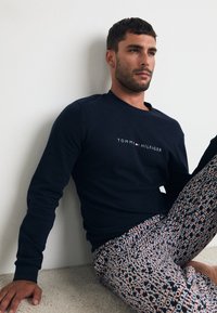 Marinegenser med "TOMMY HILFIGER"-logo, paret med mønstrede bukser som har hvite, røde og blå tekst elementer på mørk bakgrunn.