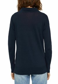 Personne aux cheveux bruns mi-longs portant une chemise marine à manches longues et un jean bleu clair, vue de dos sur fond blanc.