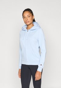 Φωτεινό γαλάζιο φούτερ με φερμουάρ, εμπρόσθια τσέπη, κουκούλα με κορδόνια και κεντημένο λογότυπο. Διαθέτει εφαρμοστά μανσέτα και απλή υφή.