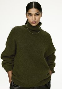 Green wool turtleneck sweater with a loose fit, rolled cuffs, and soft, fuzzy texture. Paired with a thin silver necklace, minimal design.