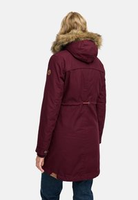Bordo parka plašč z oblogo iz krzna na kapuci, pasom za cinčenje in rjavim logotipom na rokavu. Narejen iz trpežne, teksturirane tkanine.