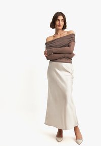 Off-the-shoulder bruine top met lange mouwen, gecombineerd met een vloeiende crème satijnen rok en puntige beigen hakken. Eenvoudig ontwerp, zachte textuur.