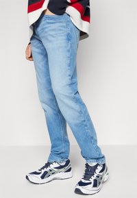 Vaqueros de mezclilla azul claro con un corte recto, combinados con zapatillas blancas y marinas con detalles en verde y un diseño texturizado.