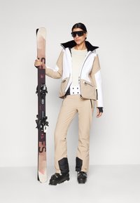 Skiausrüstung mit einer beige-weißen isolierten Jacke, passenden beigen Hosen mit schwarzen Akzenten und schwarzen Skischuhen. Hält Skier.