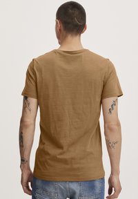 Brun kortermet t-skjorte laget av myk stoff, med rund hals og en avslappet passform. Synlige tatoveringer på armene. Enkelt design.