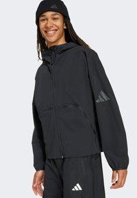 Crna kapu na jakna od laganog materijala, s prednjim zatvaračem, elastičnim manžetama i reflektirajućim detaljima. Uparena s crnim hlačama.