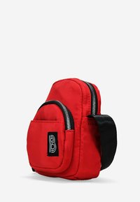 Bolsa de nylon roja con forma redondeada, que cuenta con un compartimento principal con cremallera, un bolsillo frontal y una correa negra. Detalle de logo destacado en la parte frontal.