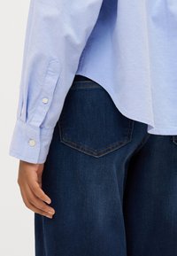 Šviesiai mėlynas marškinėlių mygtukais, su ilgomis rankovėmis ir rankogaliais, derinamas su tamsiai mėlynomis džinsomis, turinčiomis užpakalines kišenes, kurios formos primena deimantą.