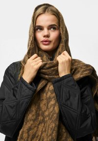 Brun mønstret tørklæde hængende over skuldrene, med en blød tekstur, og en sort quiltet jakke synlig under.