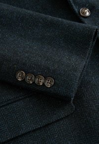 Manche de blazer en laine chevron bleu foncé avec quatre boutons marron et un bouton assorti partiellement visible sur le corps de la veste.