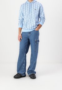 Muž v světlomodré háčkované mikině, modrých volně střižených cargo džínách s boční kapsou a černých teniskách, stojí s jednou rukou v kapse.