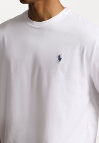 T-shirt bianca in cotone con collo rotondo, caratterizzata da un piccolo logo ricamato blu nella zona del petto sinistro. Texture morbida e design casual.