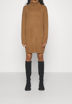 Vrouw draagt een bruin, langmouwig coltrui-jurk met geribbelde manchetten en zoom, gecombineerd met zwarte kniehoge laarzen, staand tegen een witte achtergrond.