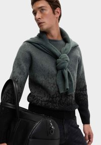 Maglione grigio a gradient con una sciarpa verde annodata drappeggiata sulle spalle, abbinato a una borsa nera texturizzata, che mette in mostra materiali morbidi.
