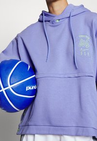 Laventelinvärinen huppari, jossa on kengurupocket ja graafinen printti sekä vihreitä yksityiskohtia. Pitelee sinistä koripalloa, jossa on valkoisia raitoja ja logo.