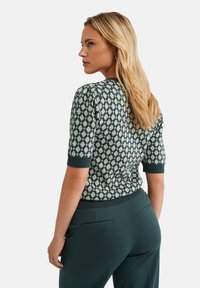 Maglione verde lavorato a maglia con maniche corte, caratterizzato da un motivo a rombi di colore chiaro, collo a coste e polsini, abbinato a pantaloni verde scuro.