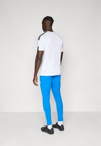 Sportstøj med en hvid T-shirt med korte ærmer og sorte striber samt blå tætsiddende bukser. Teksturede sorte sko fuldender outfittet.