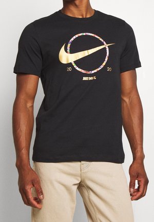 Mężczyzna w czarnej koszulce z złotym logo Nike w kolorowym okręgu, z napisem „2020” i „JUST DO IT.” z przodu, w zestawieniu z beżowymi spodniami.