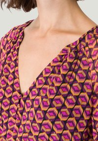 Haut en tissu violet avec un décolleté en V, arborant un motif géométrique rose, orange et doré, avec des détails froncés aux épaules.