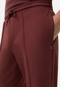 Bordeaux kleurige sportbroek met een trekkoord in de taille, zijzakken en een klein zilveren merkteken. De stof lijkt zacht en glad van textuur.
