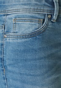 Jeans de ganga azul claro com um bolso traseiro com costura desbotada, detalhe de botão prateado e uma textura suave.