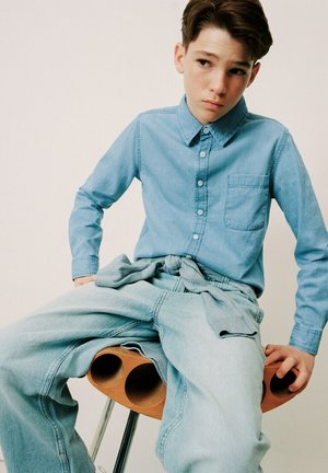 Tienerjongen met bruin haar, gekleed in een lichtblauw denim overhemd en spijkerbroek, zittend op een moderne houten kruk tegen een effen achtergrond.