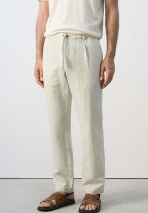 Man met lichtbeige linnen broek met trekkoord, wit overhemd en bruine sandalen met bandjes, staand tegen een egale achtergrond.