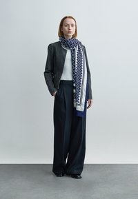 Une veste gris foncé texturée portée sur une chemise en maille blanche, associée à un pantalon noir ample. Une écharpe à motifs bleus et blancs pend sur le devant.