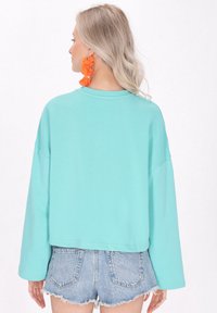 Felpa turquoise cropped con maniche lunghe, collo a costine e vestibilità rilassata, abbinata a pantaloni shorts in denim chiaro. Orecchini floreali arancioni.