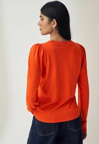Oranžs pulovers, ribots kakla izgriezums un piedurknes, pūkains plecu dizains, mīksta tekstūra, saskaņots ar tumšiem džinsu biksēm.