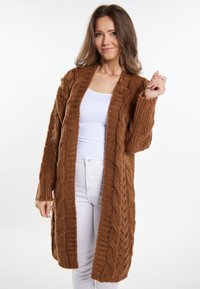 Brun strikket cardigan med kabelmønstre, åben front, lange ærmer og en hyggelig tekstur. Kombinationeret med en hvid bluse og lyse bukser.