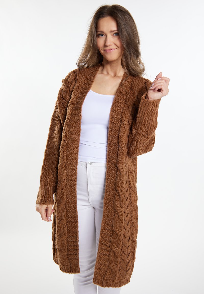 Brun strikket cardigan med kabelmønstre, åben front, lange ærmer og en hyggelig tekstur. Kombinationeret med en hvid bluse og lyse bukser.