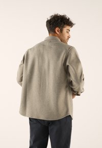 Gray wool-blend shirt with a pointed collar, visible seams, and a curved hem. Long sleeves with rolled cuffs, displayed from the back.
