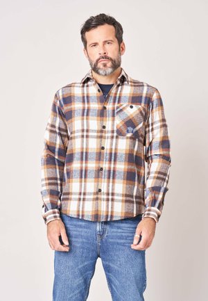 Volwassen man met baard die een bruin, blauw en wit geruit overhemd met knopen en blauwe spijkerbroek draagt, staand tegen een effen lichte achtergrond.
