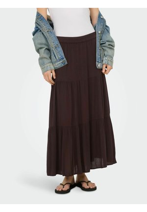 Kobieta ubrana w warstwową ciemnobrązową długą spódnicę, biały top, jeansową kurtkę i czarne klapki, stojąca na jednolitym tle.